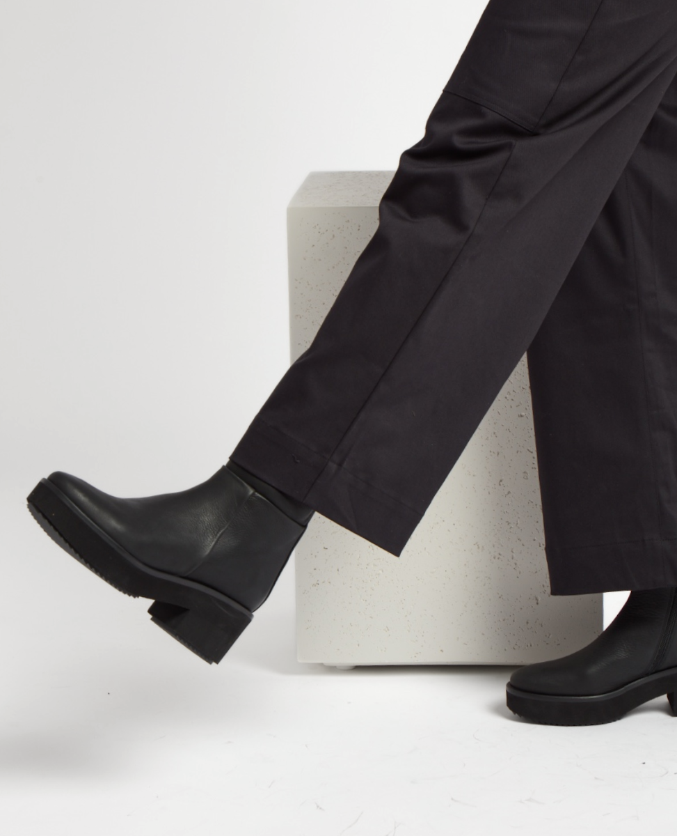 A pair of sleek black leather pleatform boots with a smooth texture, featuring a low block heel and a rounded toe, worn with dark pants.