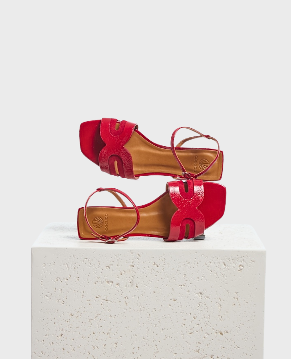 A pair of vibrant red sandals with a glossy finish, featuring a square toe, intricate cut-out design on the straps, and a flat sole, displayed on a textured white pedestal.