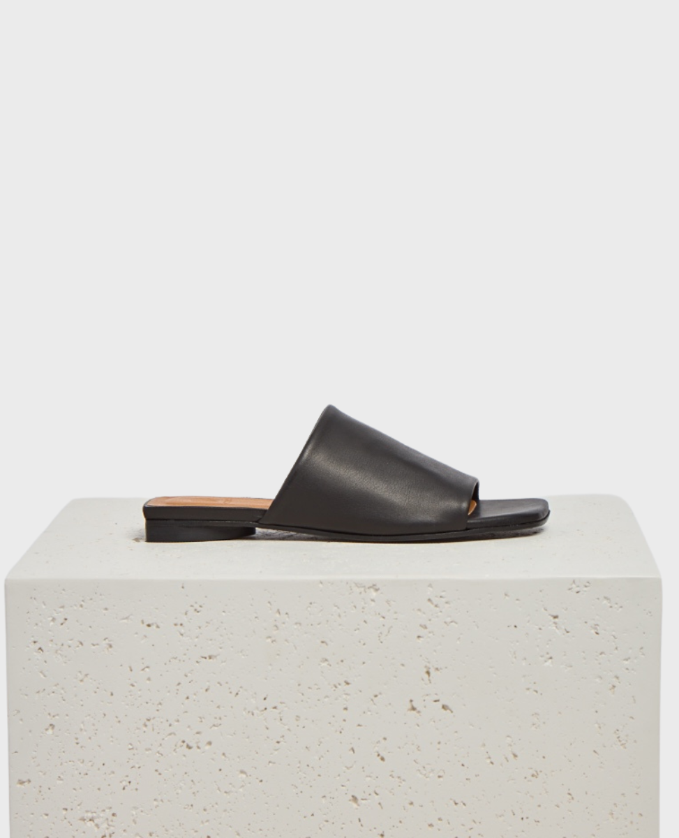 A sleek black leather slide sandal with a minimalist design, featuring a wide strap across the foot, an open toe, a flat sole, and a subtle heel.