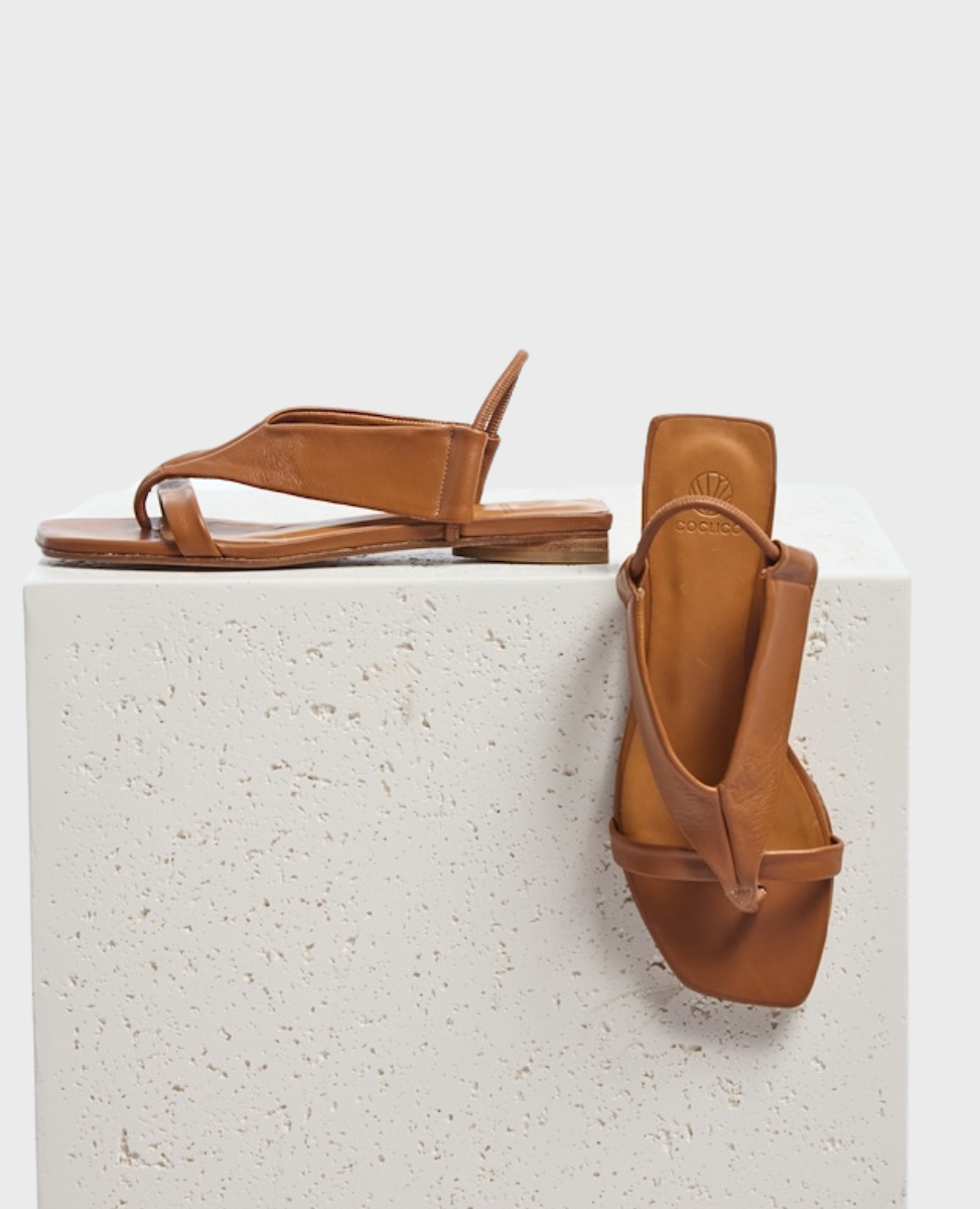 Brown leather sandals with a minimalist design feature a thong strap and a square toe, resting on a textured white surface.