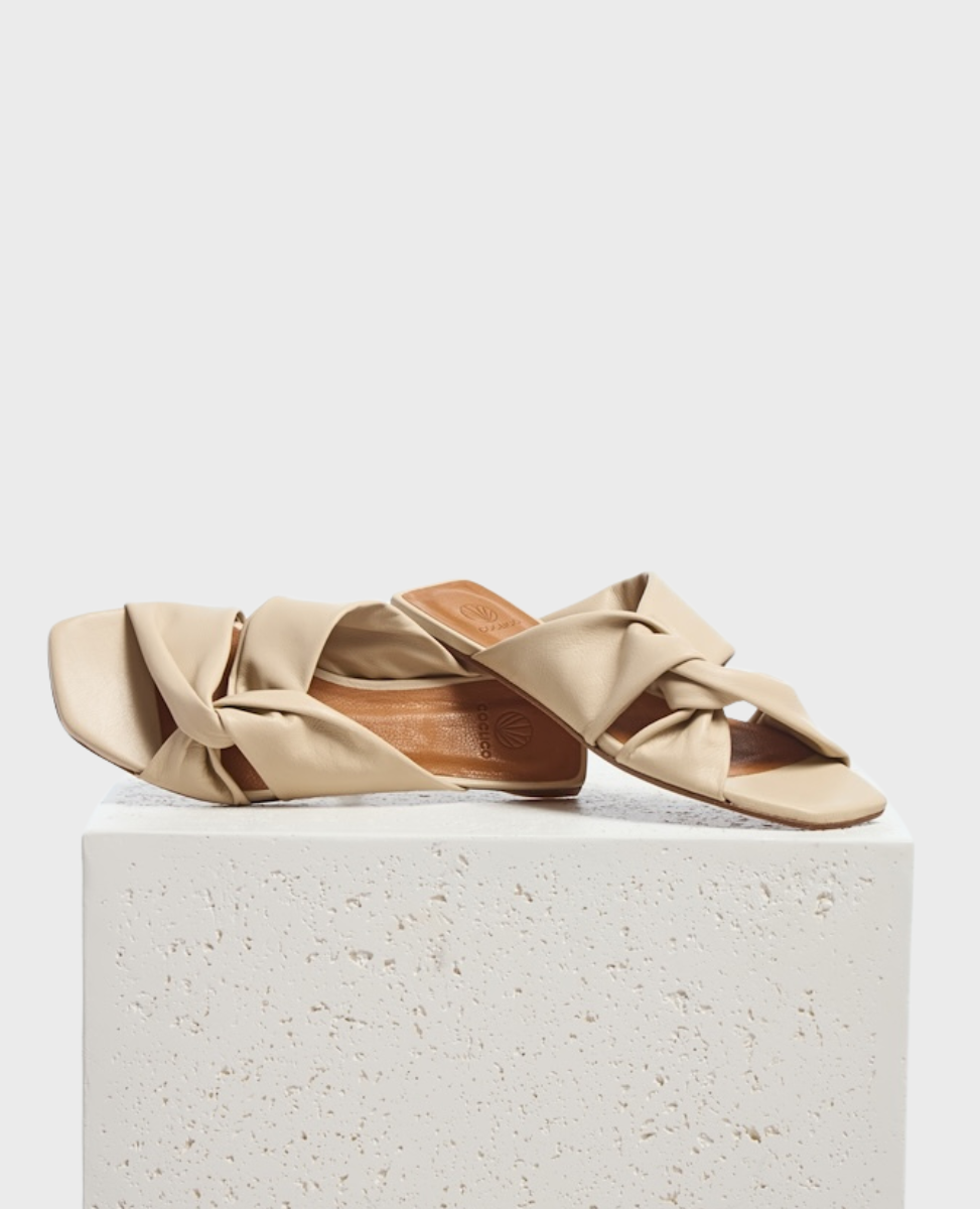 Beige sandals with a twisted strap design, featuring a square toe and flat sole, crafted from smooth, soft material.