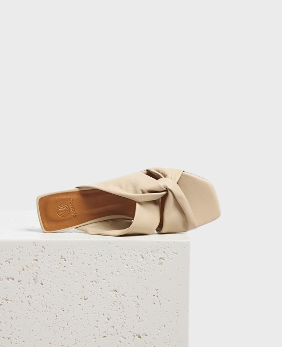 A beige, square-toed slide sandal with a twisted, soft leather upper and a flat sole, featuring a minimalist design and a subtle logo on the insole.