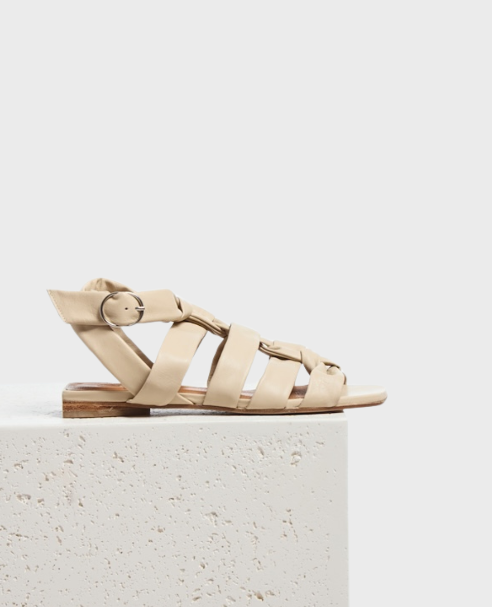 A beige gladiator sandal with multiple wide leather straps, a small buckle on the side, a flat sole, and a minimalist design.