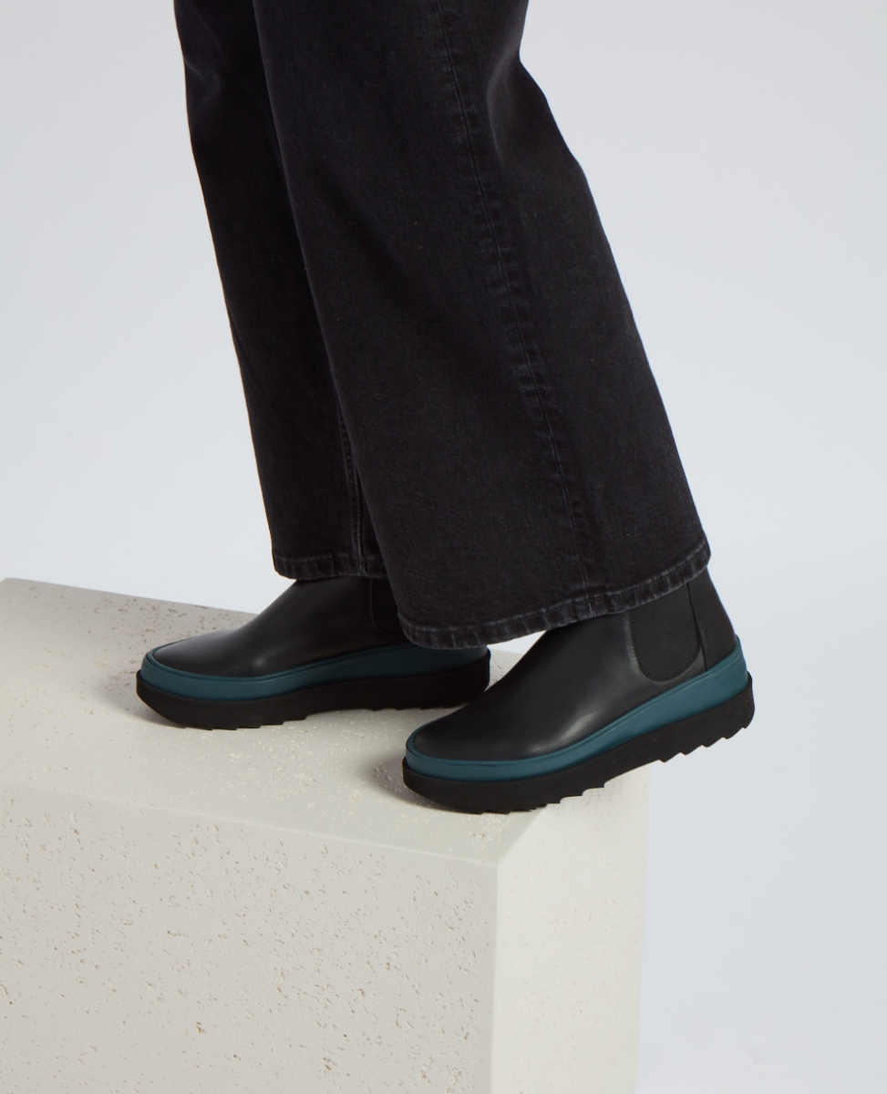 Black leather ankle boots with a smooth texture, featuring a teal rubber sole and elastic side panels, worn by a person in dark pants standing on a light-colored block.