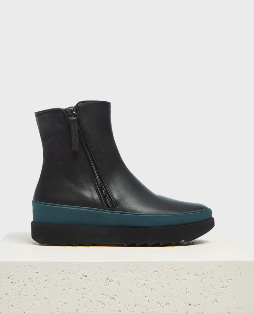 A sleek, black ankle boot with a smooth leather finish, featuring shearling lining, a side zipper, a contrasting teal rubber midsole, and a black EVA sole with a ridged texture, displayed on a white textured pedestal.
