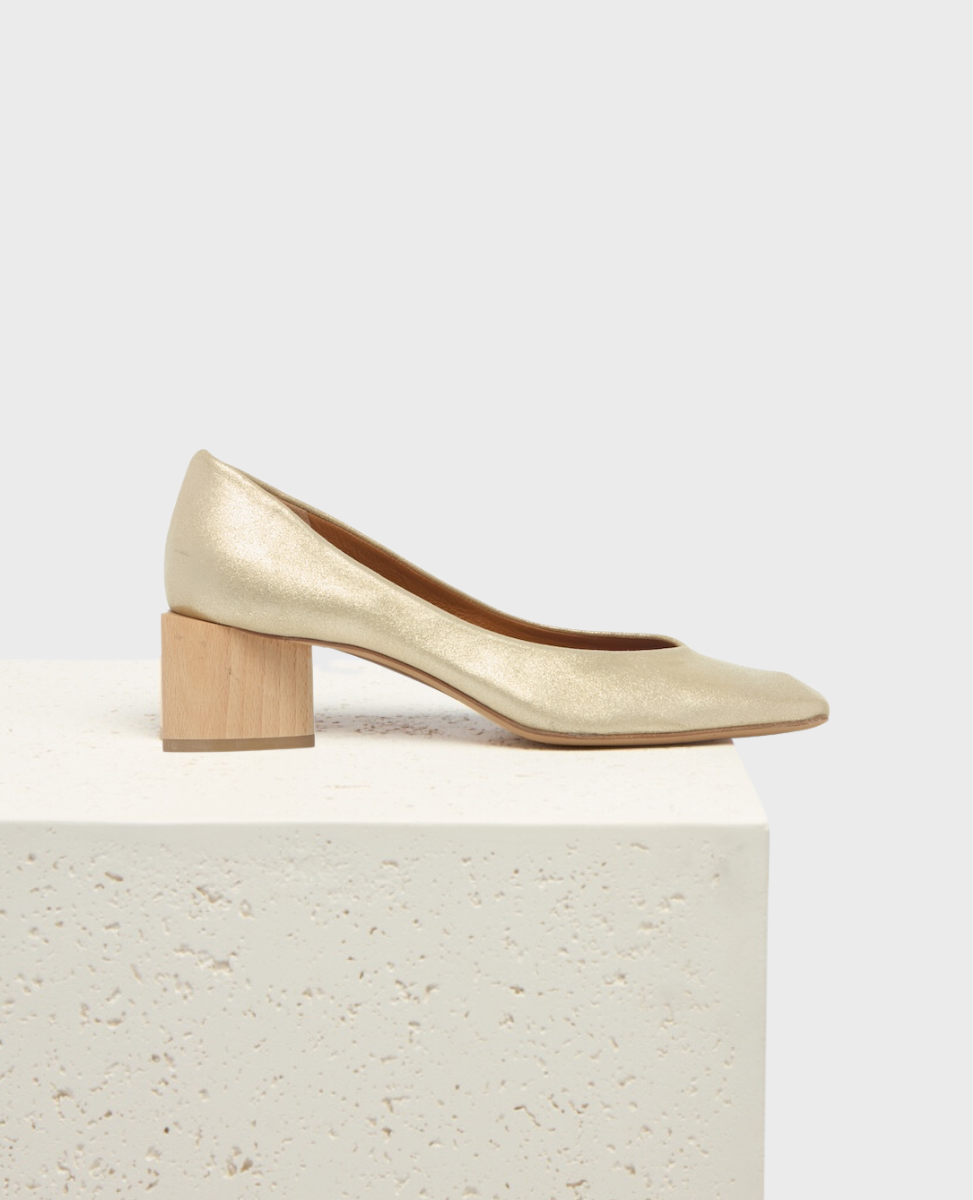 A gold, metallic-textured pump with a sleek, pointed toe and a distinctive wooden block heel, resting on a textured white surface.