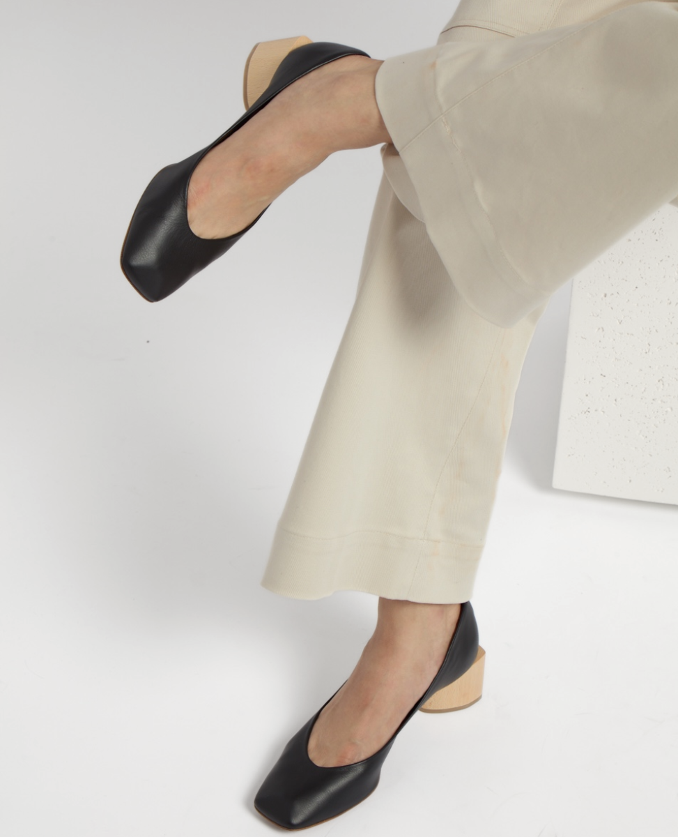 A pair of black leather shoes with a square toe and a contrasting light wooden block heel, worn with cream-colored pants.
