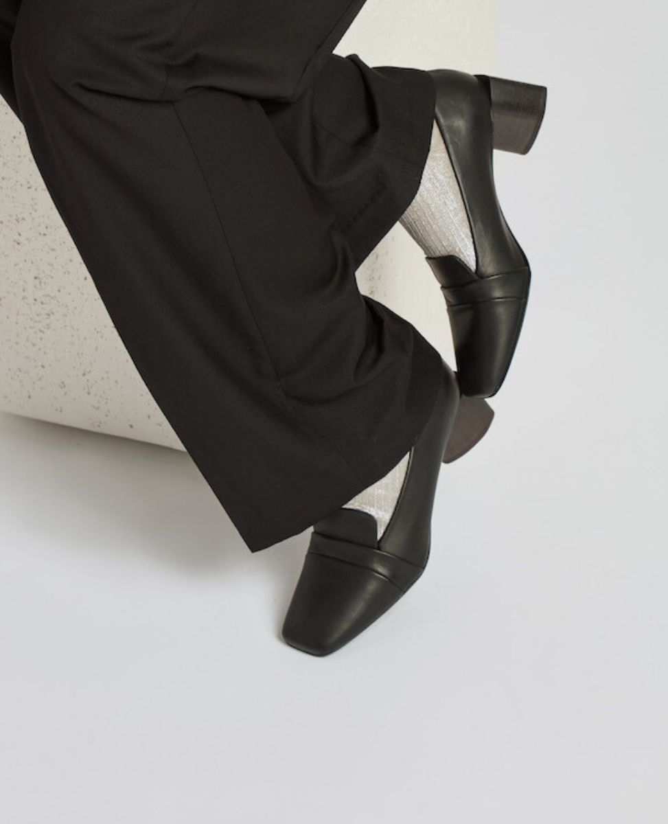Black leather loafers with a low block heel, featuring a sleek, smooth finish and a classic, minimalist design, worn with white socks and black pants.
