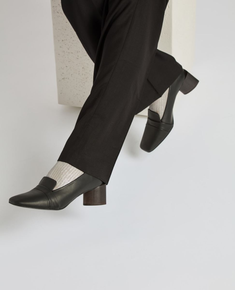 A pair of sleek, black leather loafers with a low, chunky heel, worn with black pants and light-colored socks, featuring a smooth texture and a minimalist design.