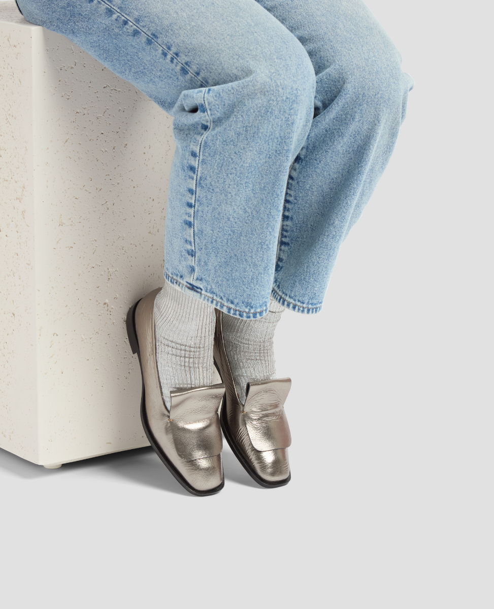 Person wearing silver loafers and light blue jeans sitting on a white stool.