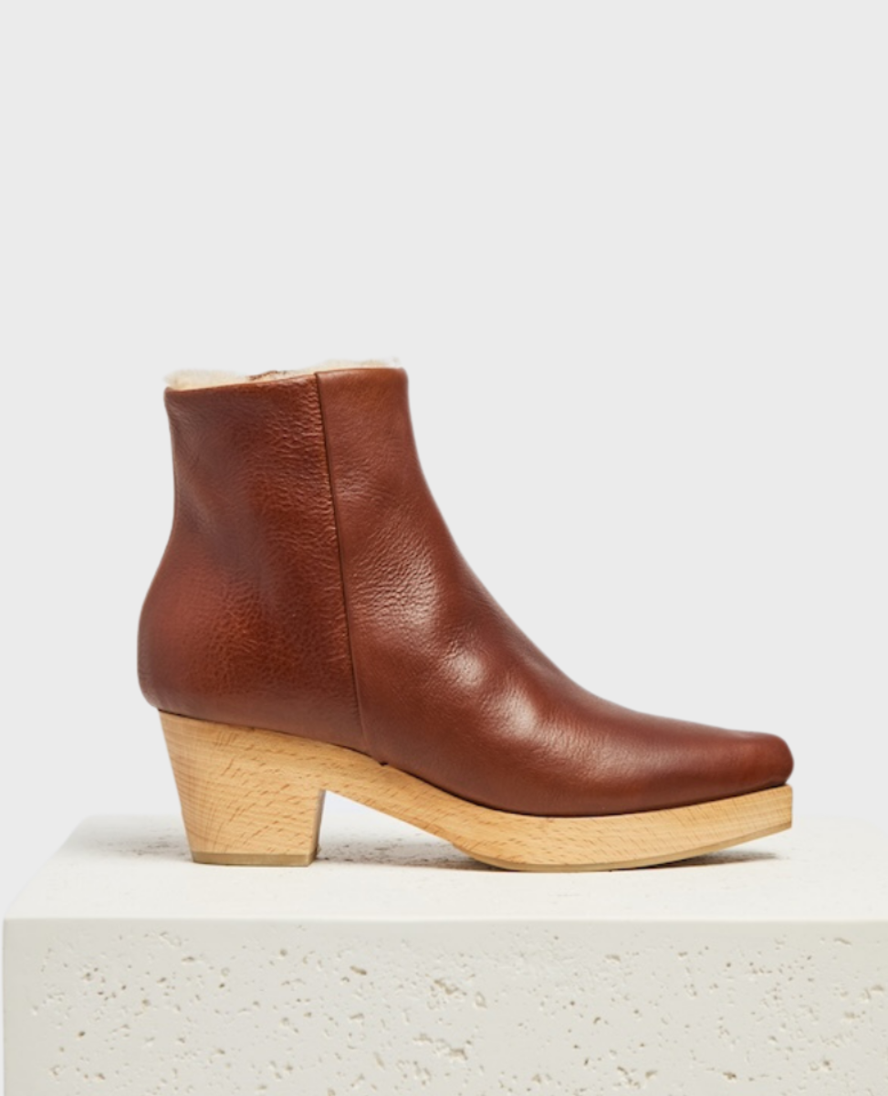 A brown leather ankle clog boot with a smooth texture, featuring a snip toe, a side zipper, a light wooden block heel, and a shearling, cream-colored inner lining.