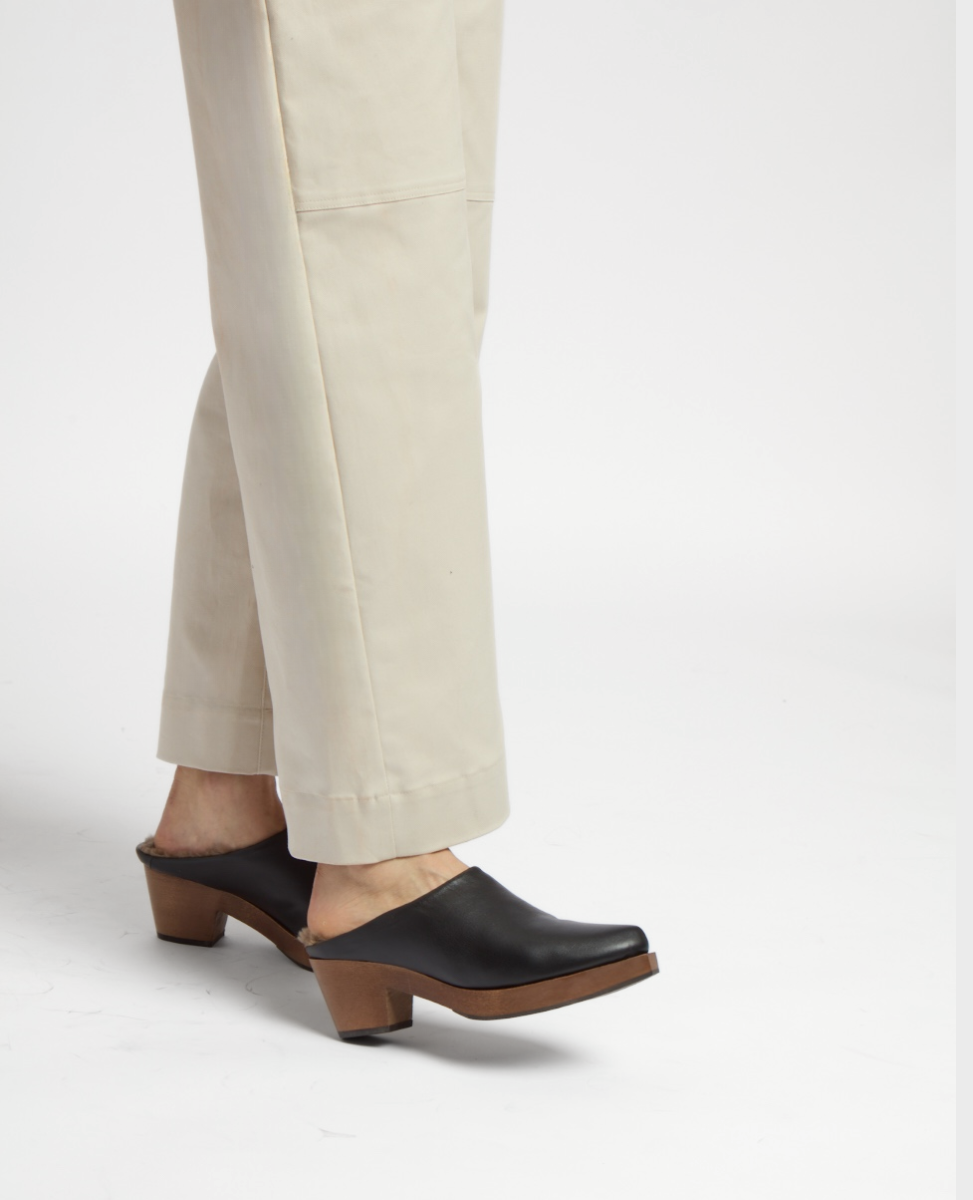 A pair of black leather clogs with a smooth texture, featuring a wooden sole and a chunky heel, worn with beige pants.
