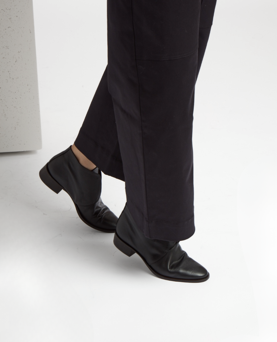 A pair of sleek, black leather ankle boots with a smooth texture, rounded toes, and low heels, worn with dark, straight-leg pants.