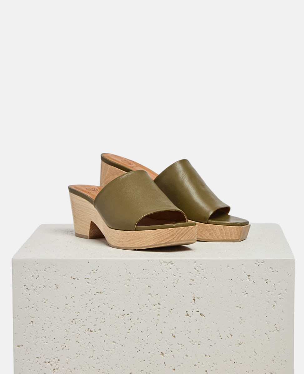 A stylish clog sandal featuring an olive green leather upper, an open toe design, and a light wooden block heel and platform. On a plinth