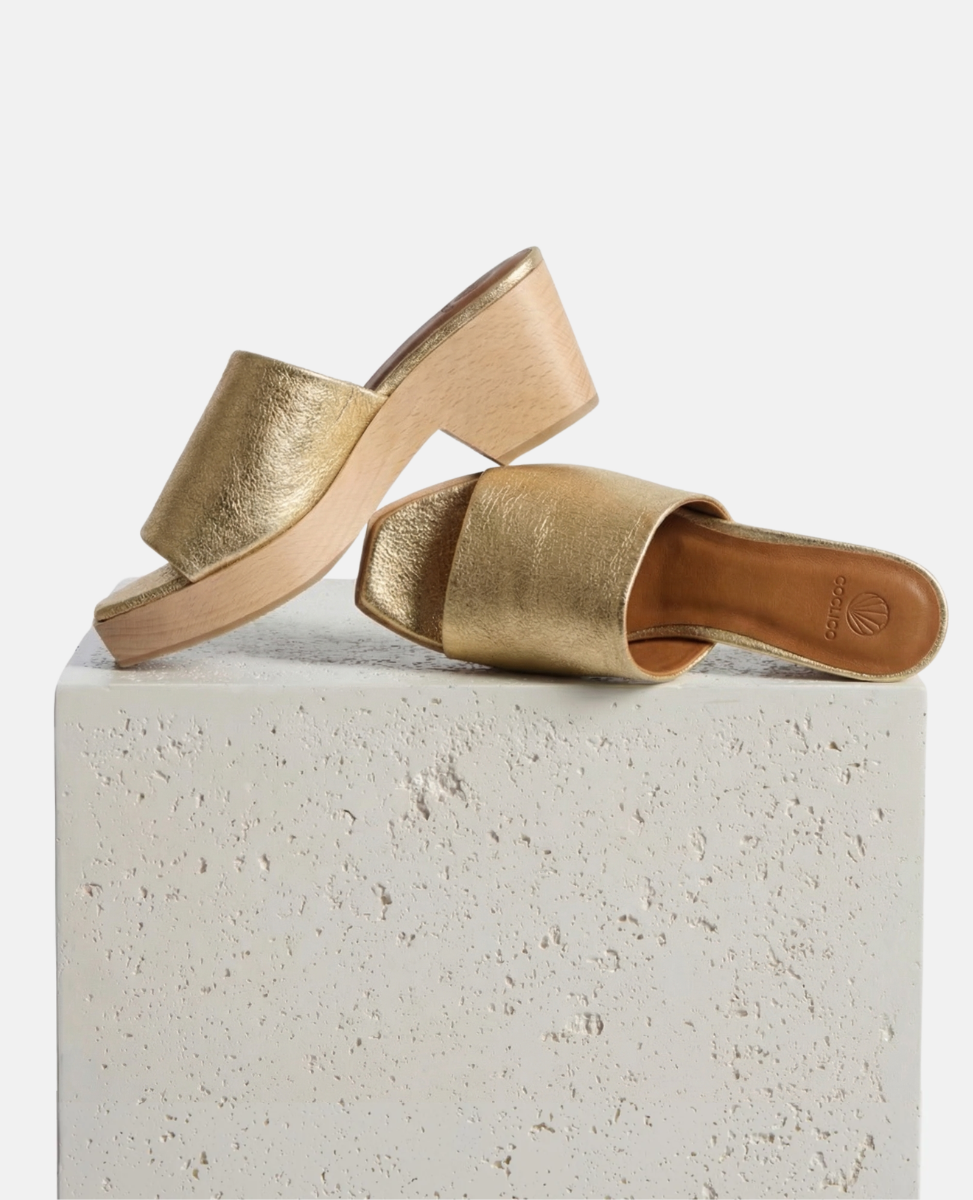 Gold leather women's wood clog sandals on a plinth.