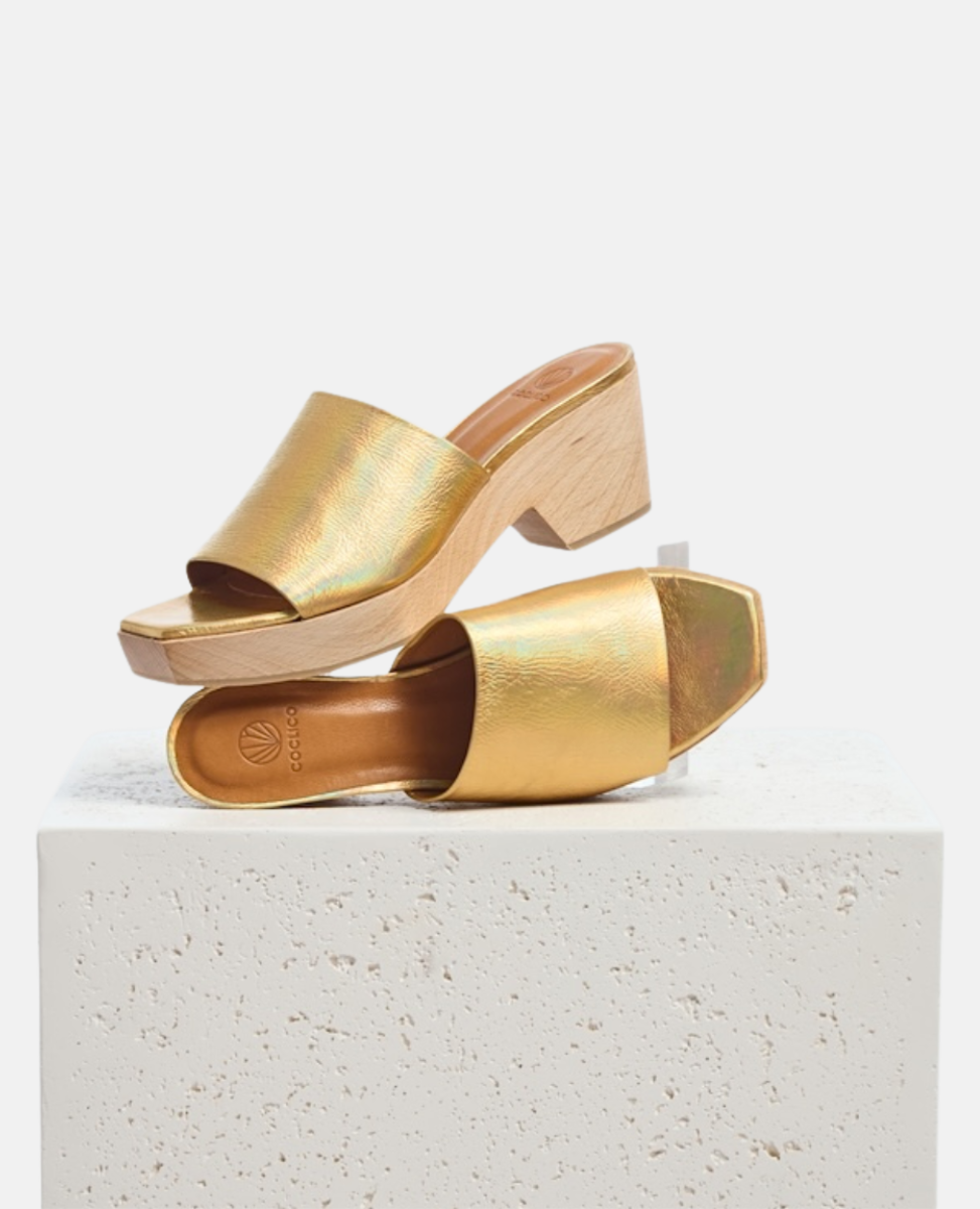 A pair of open-toe, gold iridescent slide sandals with a chunky wooden block heel and a square toe design, featuring a smooth, shiny texture and a minimalist style.