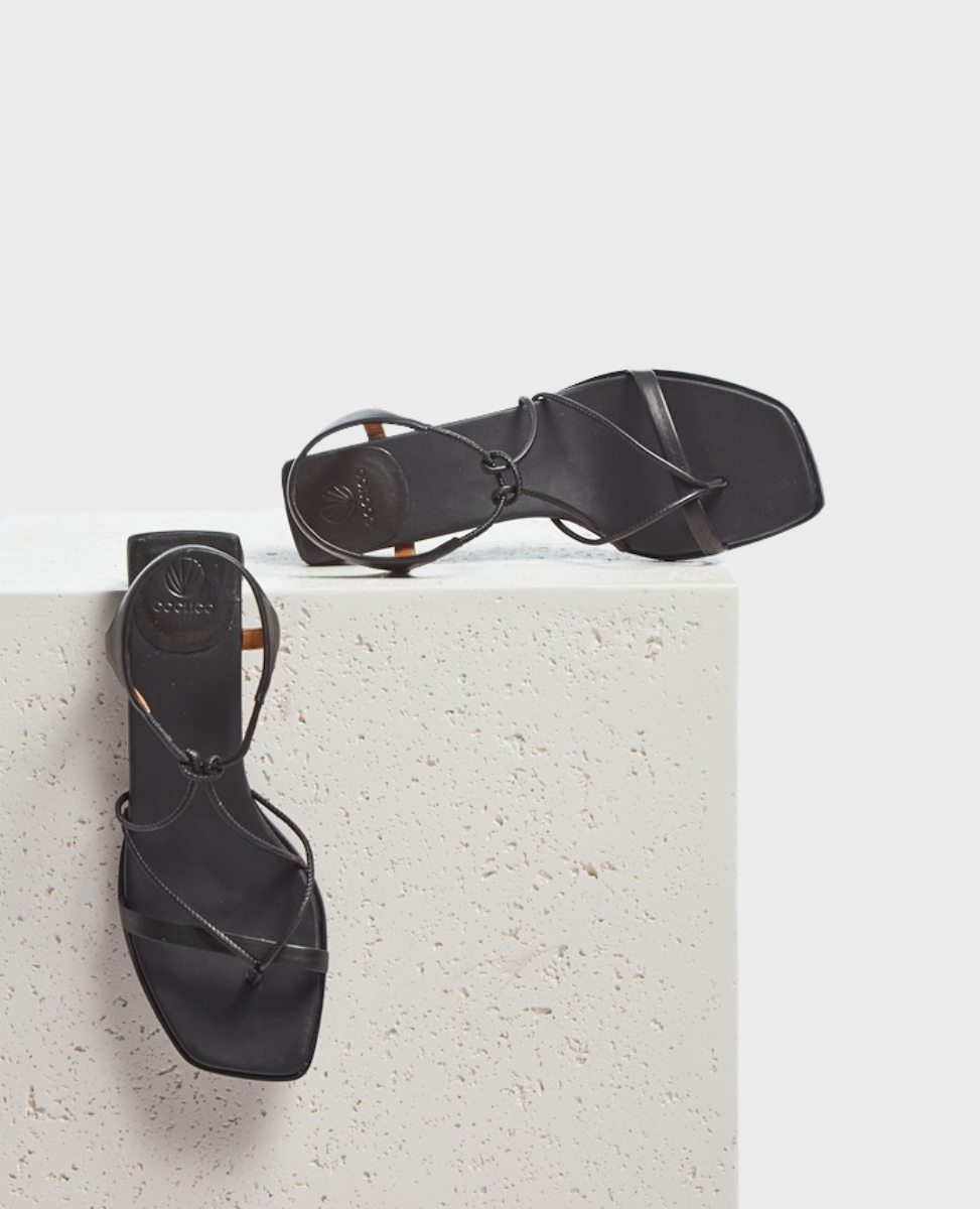 A pair of sleek, black sandals with thin, crisscrossing straps and a square toe design, featuring a smooth leather texture and minimalist aesthetic.