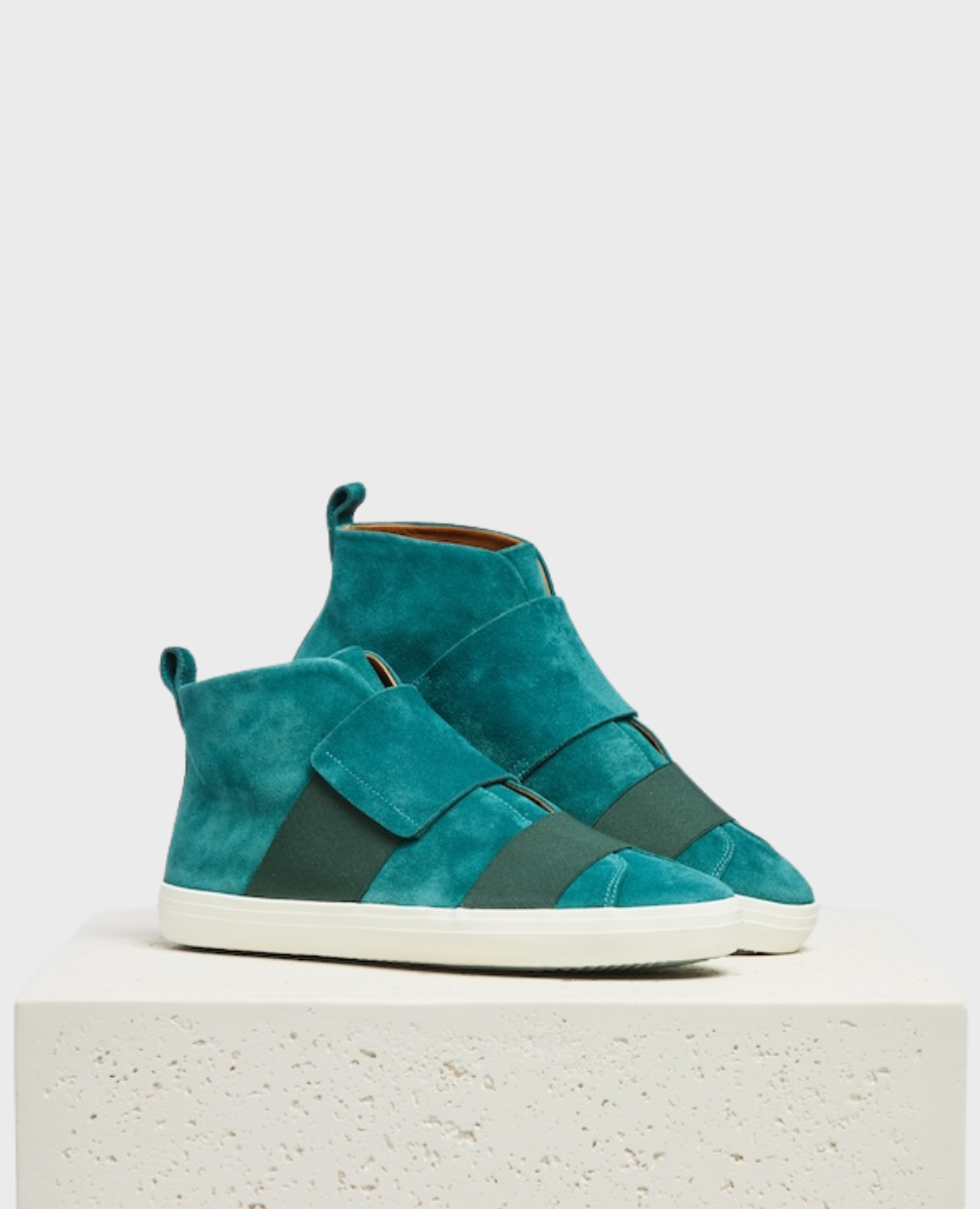 A pair of teal suede high-top sneakers with white rubber soles, featuring wide tonal elastic straps across the front and a pull tab at the heel, displayed on a textured white pedestal.