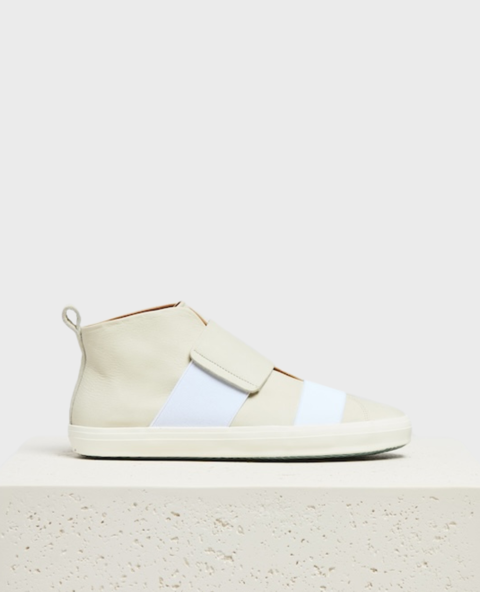 A minimalist, off-white high-top sneaker with a smooth leather texture, featuring a wide, white elastic strap across the front, a pull tab at the heel, and a white rubber sole.