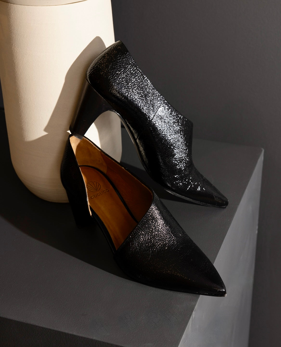 A pair of sleek, black, pointed-toe high heels with a glossy, textured finish, medium-height block heels, and a smooth, tan inner lining.