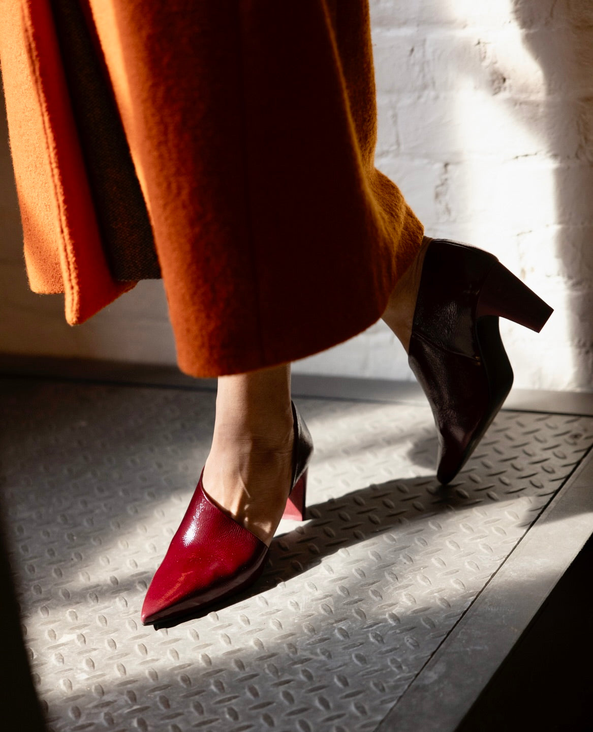 A person wearing a long orange coat and stylish cordovan patent leather high-heeled shoes walks on a textured floor illuminated by sunlight.