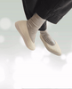 A pair of beige slip-on shoes with a smooth, matte finish, rounded toes, and thick, white rubber soles, worn with light gray socks and black pants. 12
