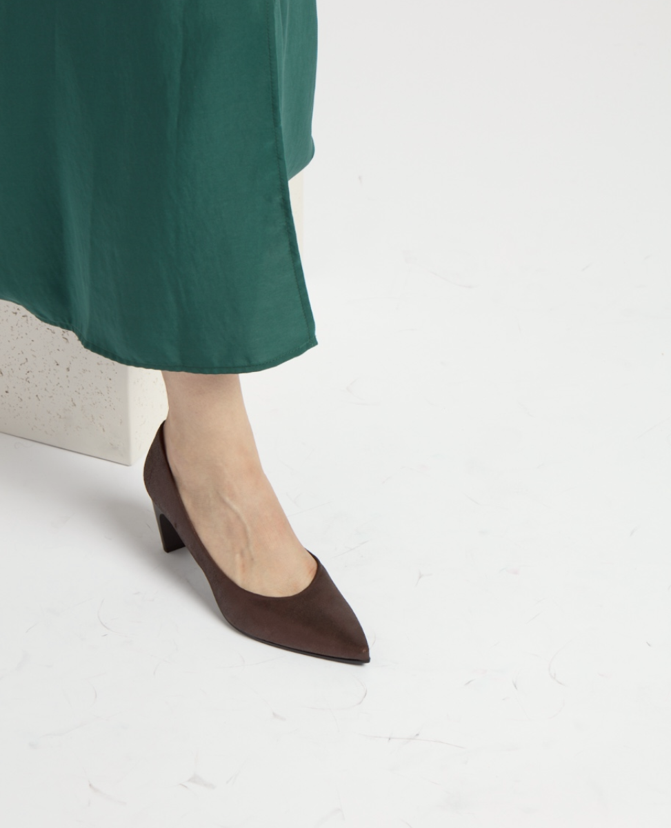 A brown metallic, pointed-toe, mid-heel shoe with a smooth leather texture is worn on a foot, partially visible beneath a flowing green fabric.