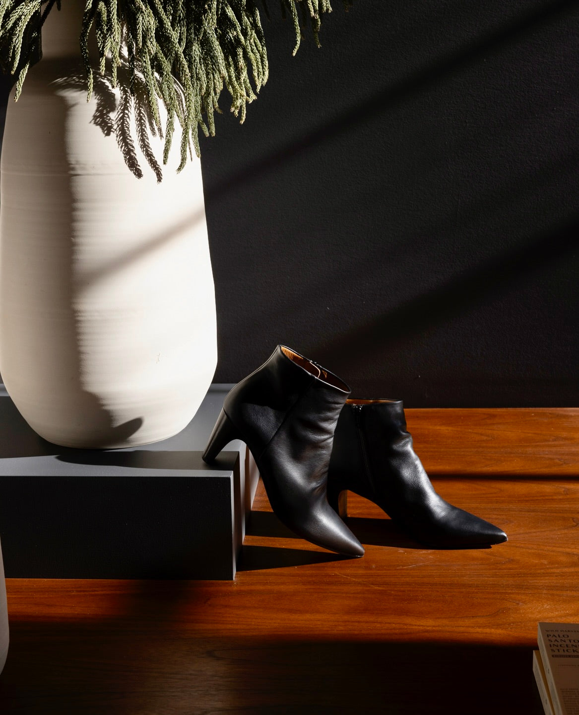 A pair of sleek, black leather ankle boots with pointed toes and solid wood heels is placed on a wooden surface next to a tall, white ceramic vase with green foliage.