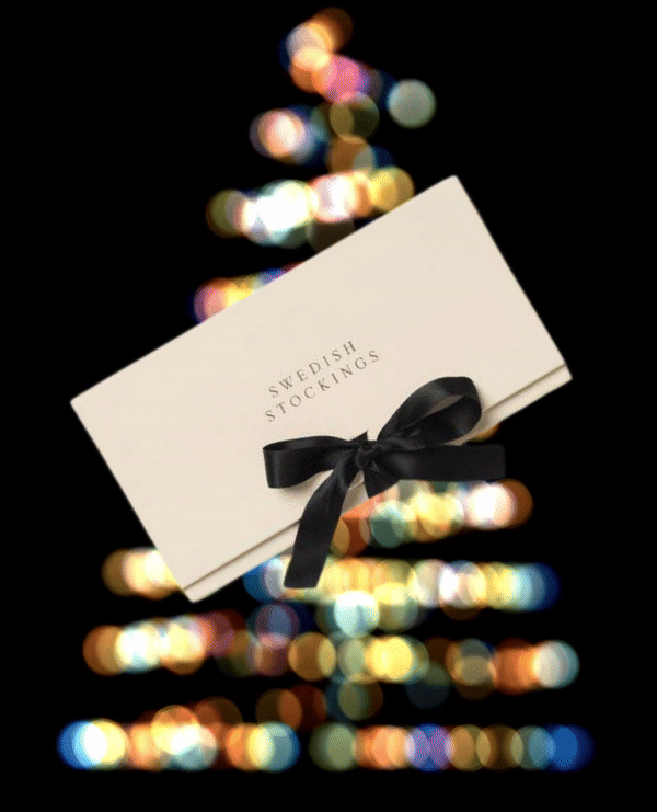 A cream-colored rectangular envelope with "SWEDISH STOCKINGS" printed in black, adorned with a sleek black ribbon tied in a bow, set against a blurred background of colorful bokeh lights resembling a Christmas tree.