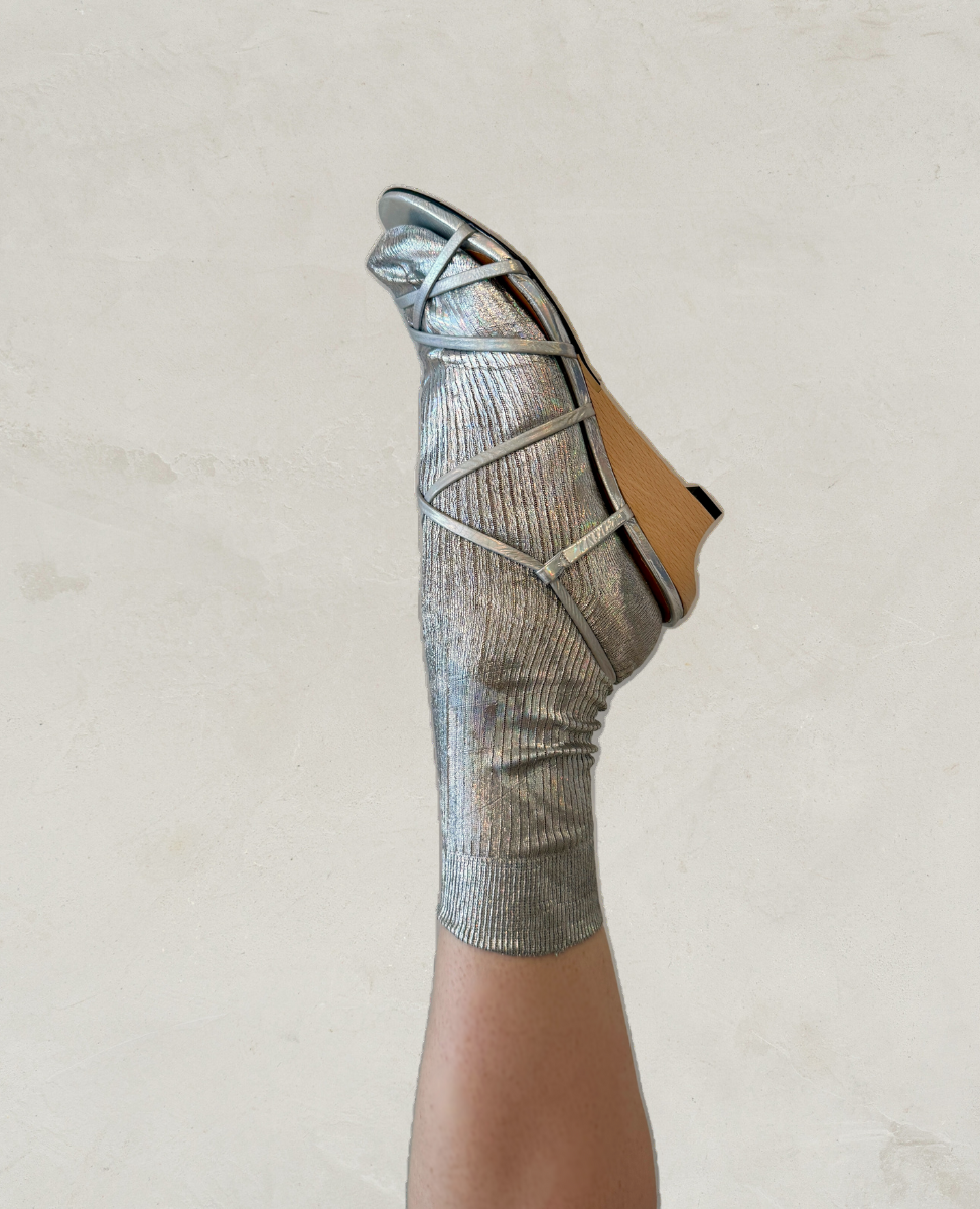 A metallic silver sock with a ribbed texture is worn with a wooden wedge sandal featuring crisscrossing silver straps.