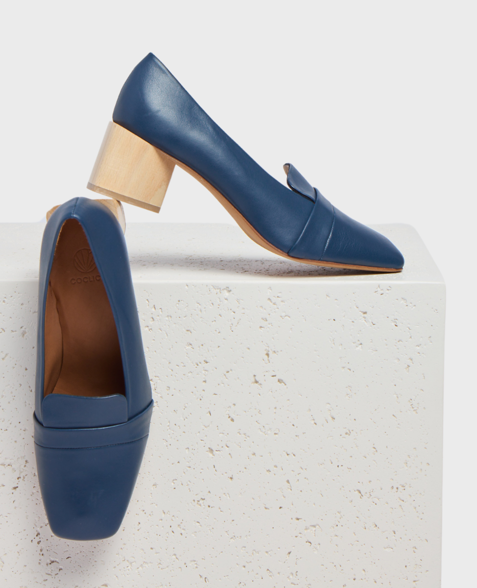A pair of elegant blue leather loafers with a sleek, pointed toe and a distinctive light wooden block heel, featuring a smooth texture and a subtle seam detail across the upper.