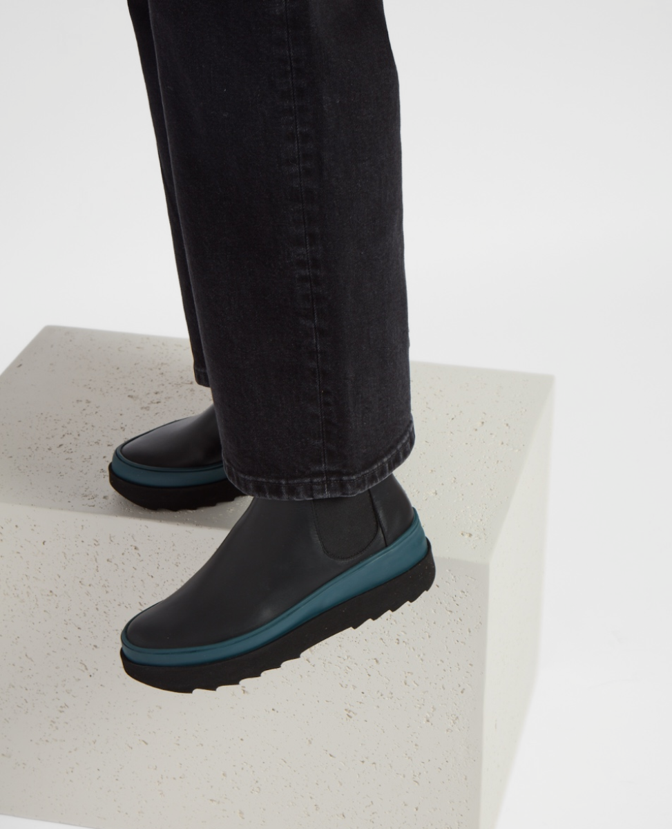 A sleek, black ankle boot with a smooth leather texture, featuring a distinctive teal sole and elastic side panels, worn with dark pants.