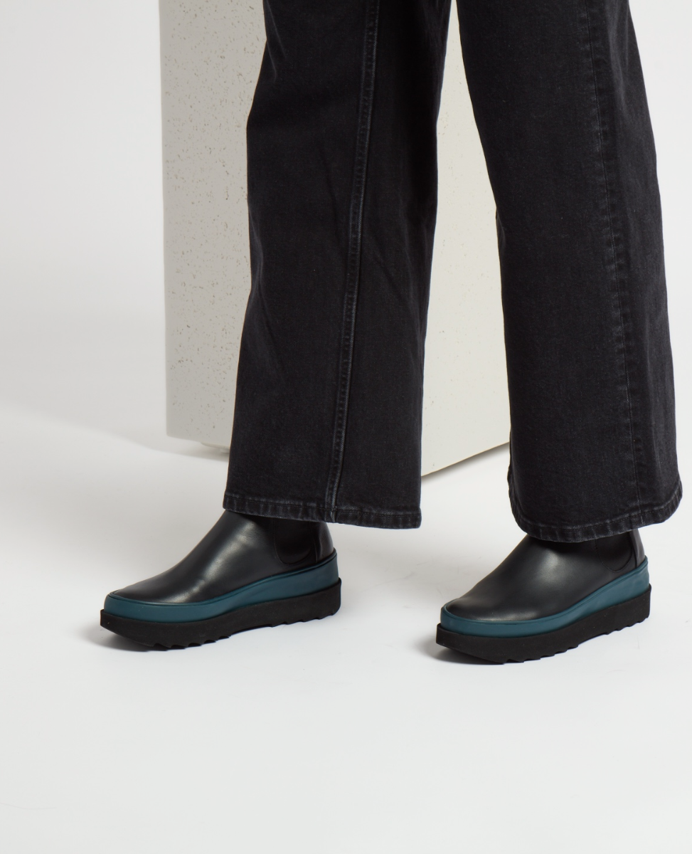 A pair of sleek, black leather slip-on shoes with a smooth texture, featuring a distinctive teal rubber sole and a rounded toe, worn with black denim pants.