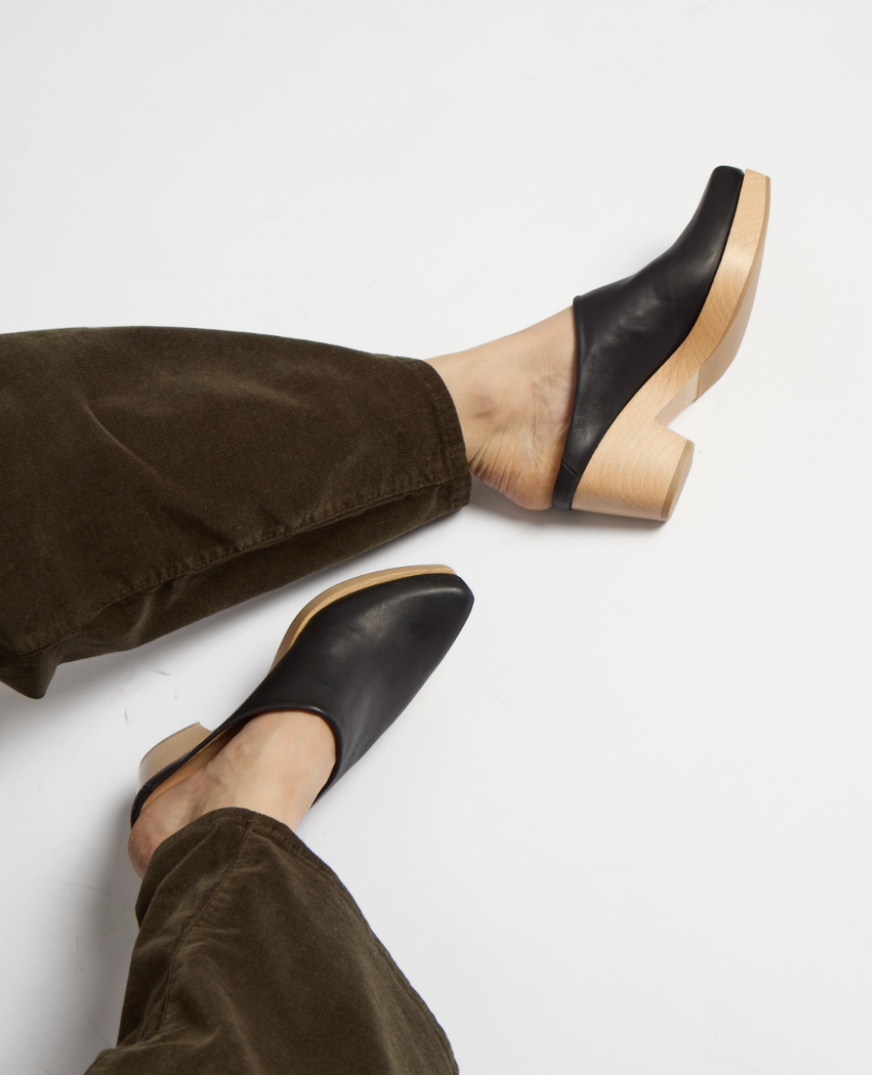 Black leather clogs with a smooth texture, featuring a chunky, light wooden heel and a closed, slightly squared toe design.