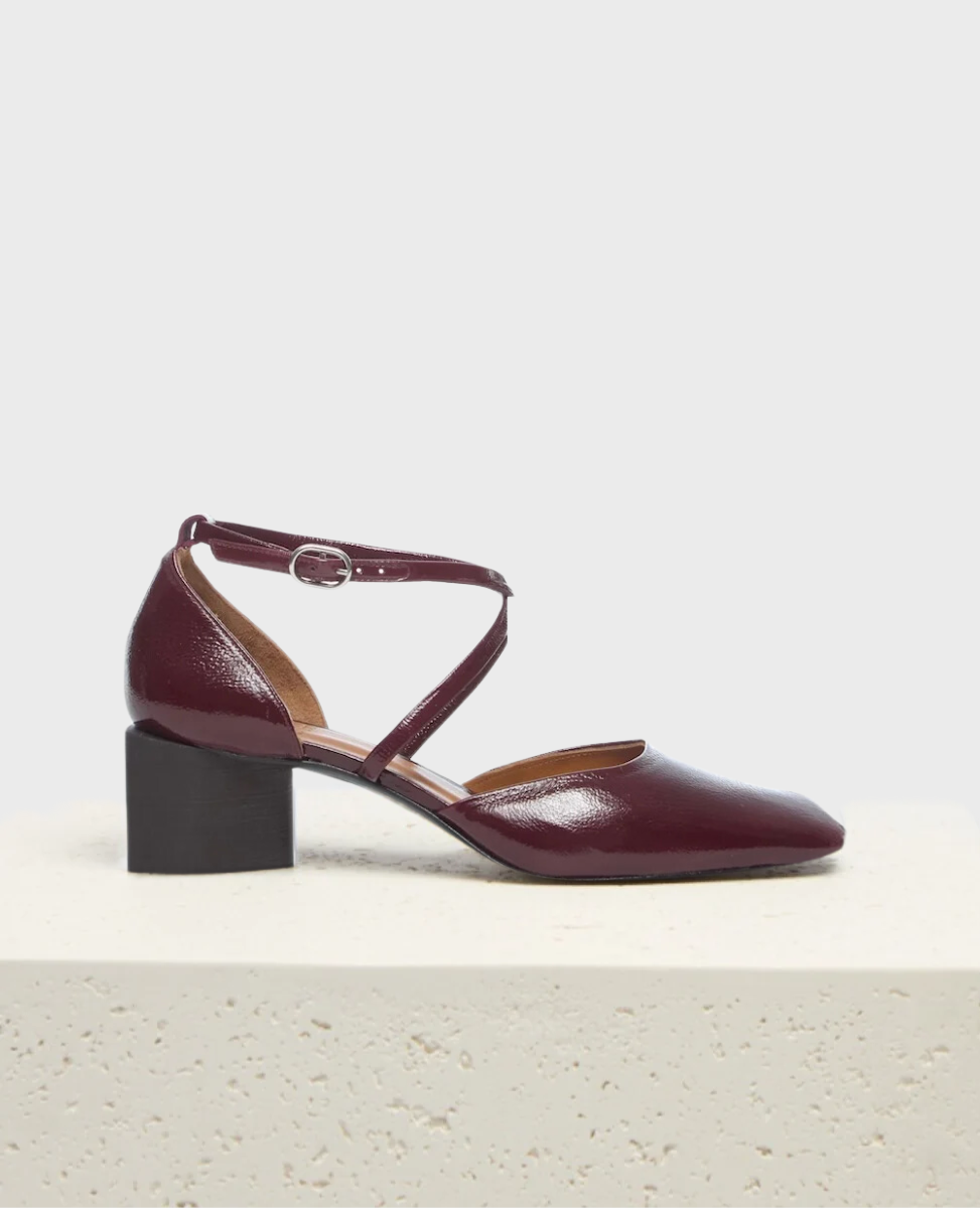 A burgundy patent leather shoe with a low block heel, pointed toe, and a crisscross ankle strap featuring a small silver buckle.