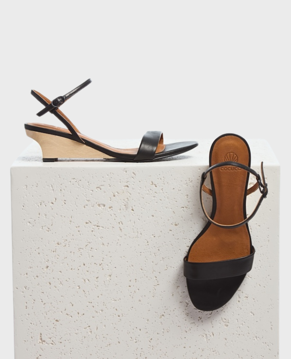 A pair of elegant black leather sandals featuring a minimalist design with a thin ankle strap, a single toe strap, and a low, light wooden wedge heel, displayed on a textured white block.