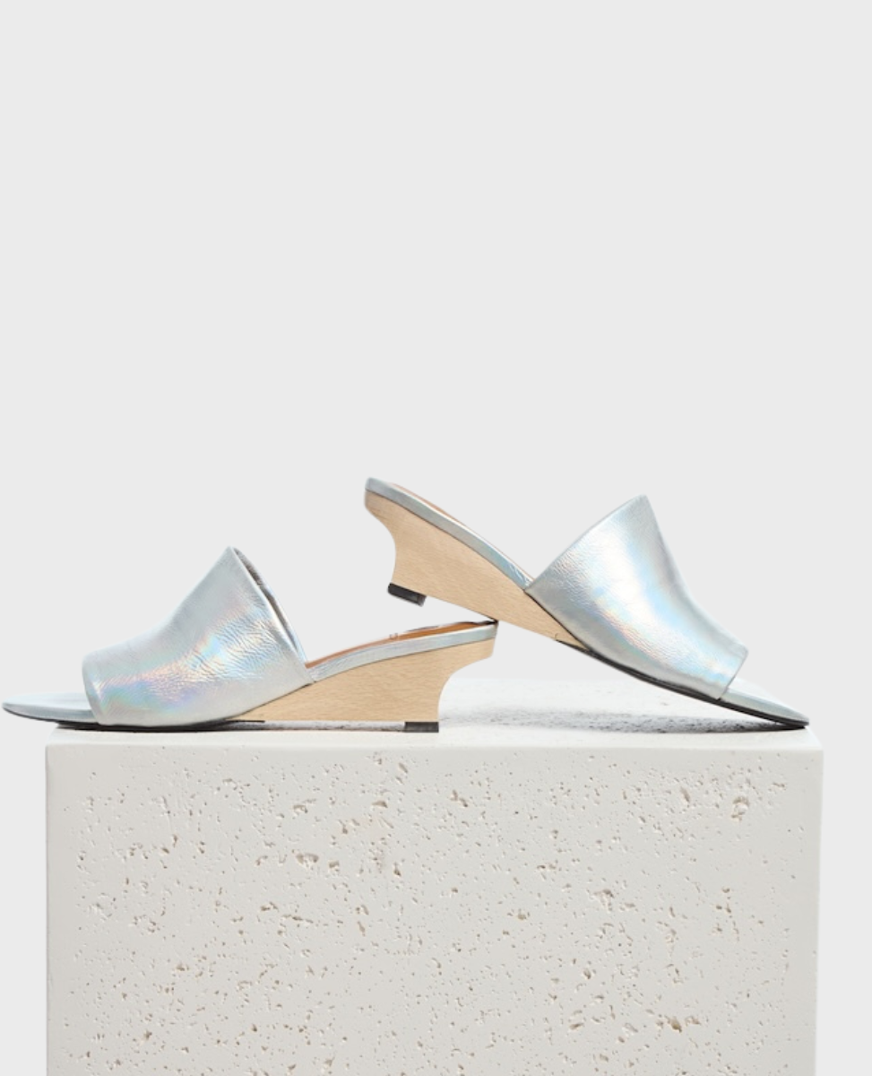 Silver, iridescent open-toe mules with a sleek, shiny finish, featuring a low, angular wooden wedge heel and a minimalist design.