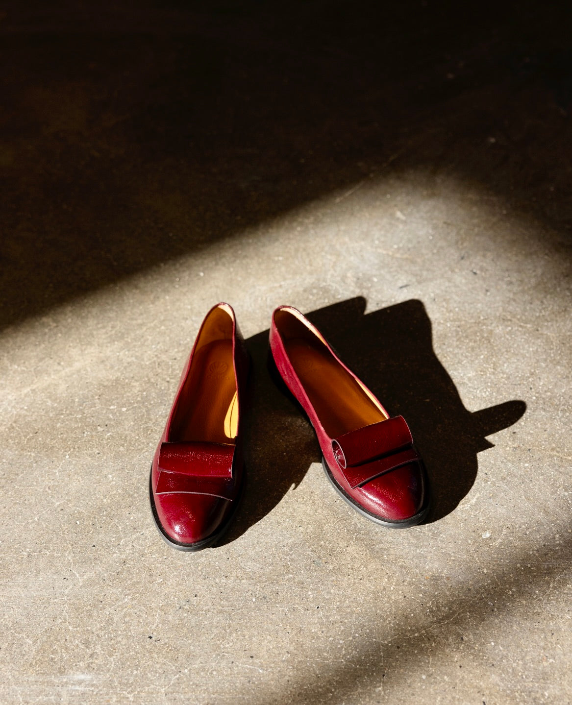 A pair of burgundy leather loafers is placed on a concrete floor, illuminated by a beam of sunlight.