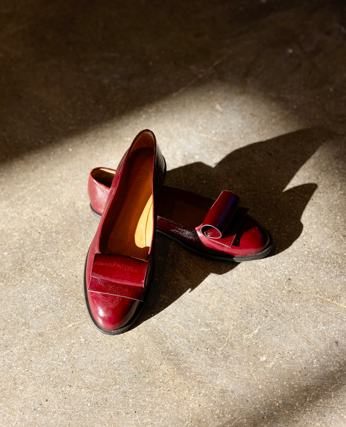 A pair of burgundy leather loafers with an architectural loop detail is placed on a concrete floor, illuminated by sunlight.