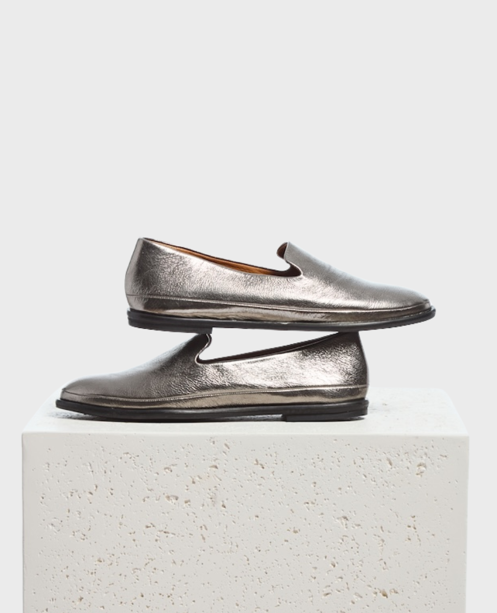A pair of metallic silver loafers with a sleek, smooth texture, featuring a low heel and a simple slip-on design, stacked on a textured white block.