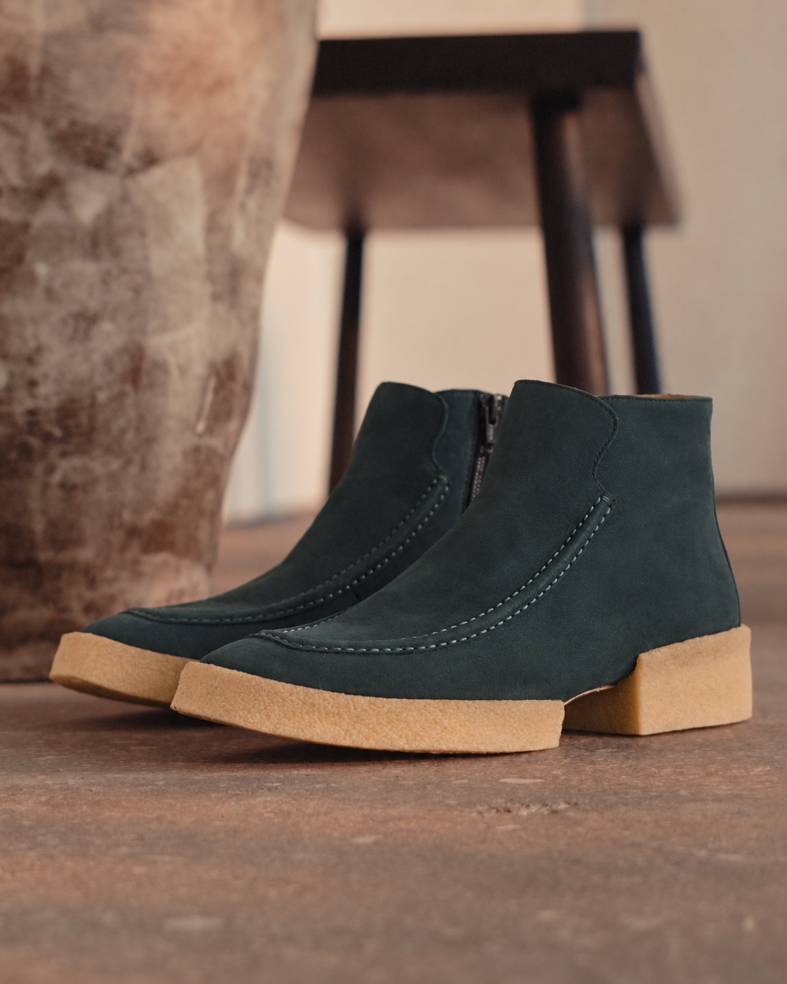 A pair of deep sea suede ankle boots with natural crepe rubber soles, displayed on a rustic wooden floor next to a large stone vase.