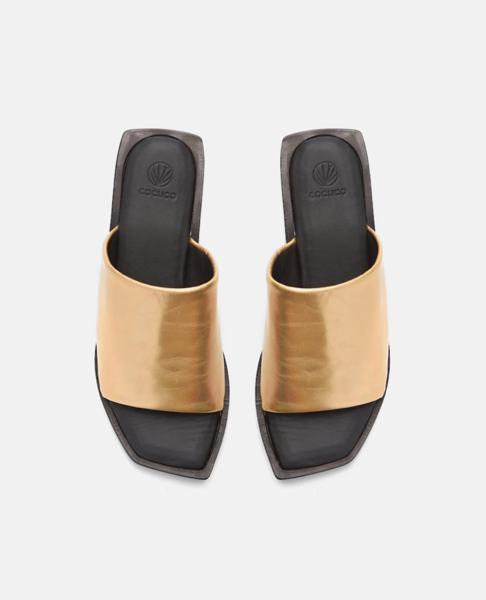 A pair of open-toed, square-heeled sandals featuring a wide, metallic gold strap across the top, with a sleek black footbed and a subtle logo embossed on the insole.
