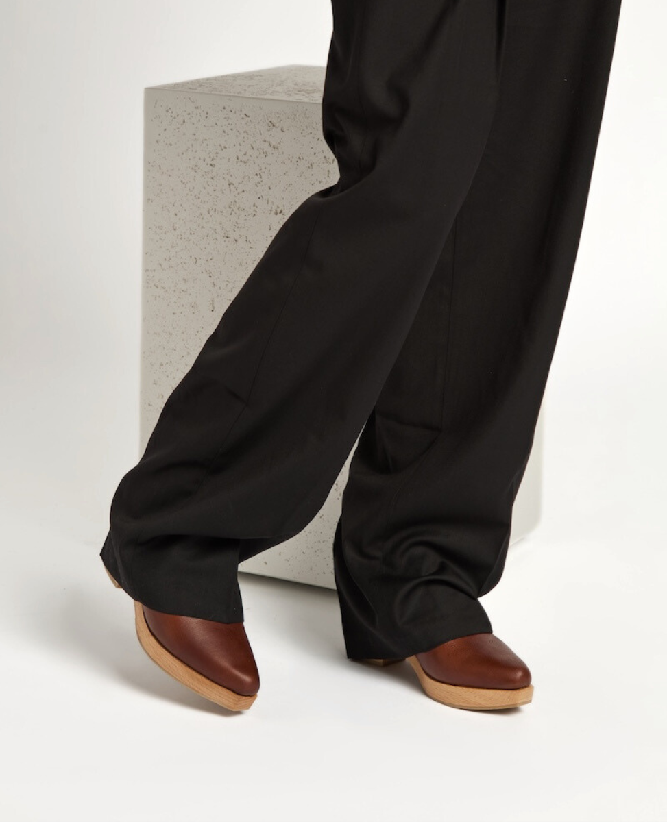 A pair of brown leather clogs with a wooden sole, featuring a sleek, snipped toe design, worn with black, wide-legged pants.