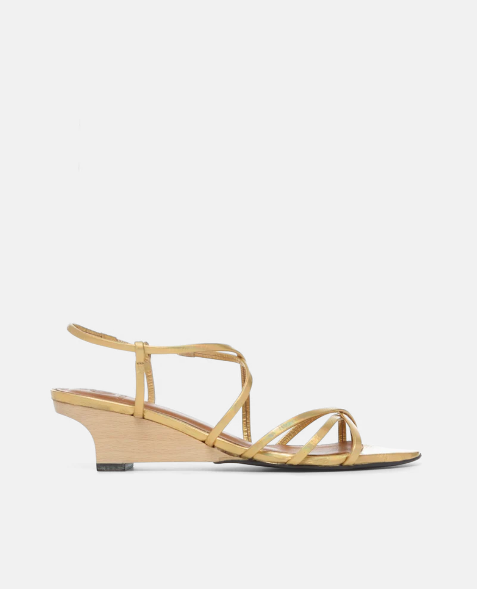 A sleek, gold sandal with thin, crisscrossing leather straps, a low wooden wedge heel, and a minimalist design.