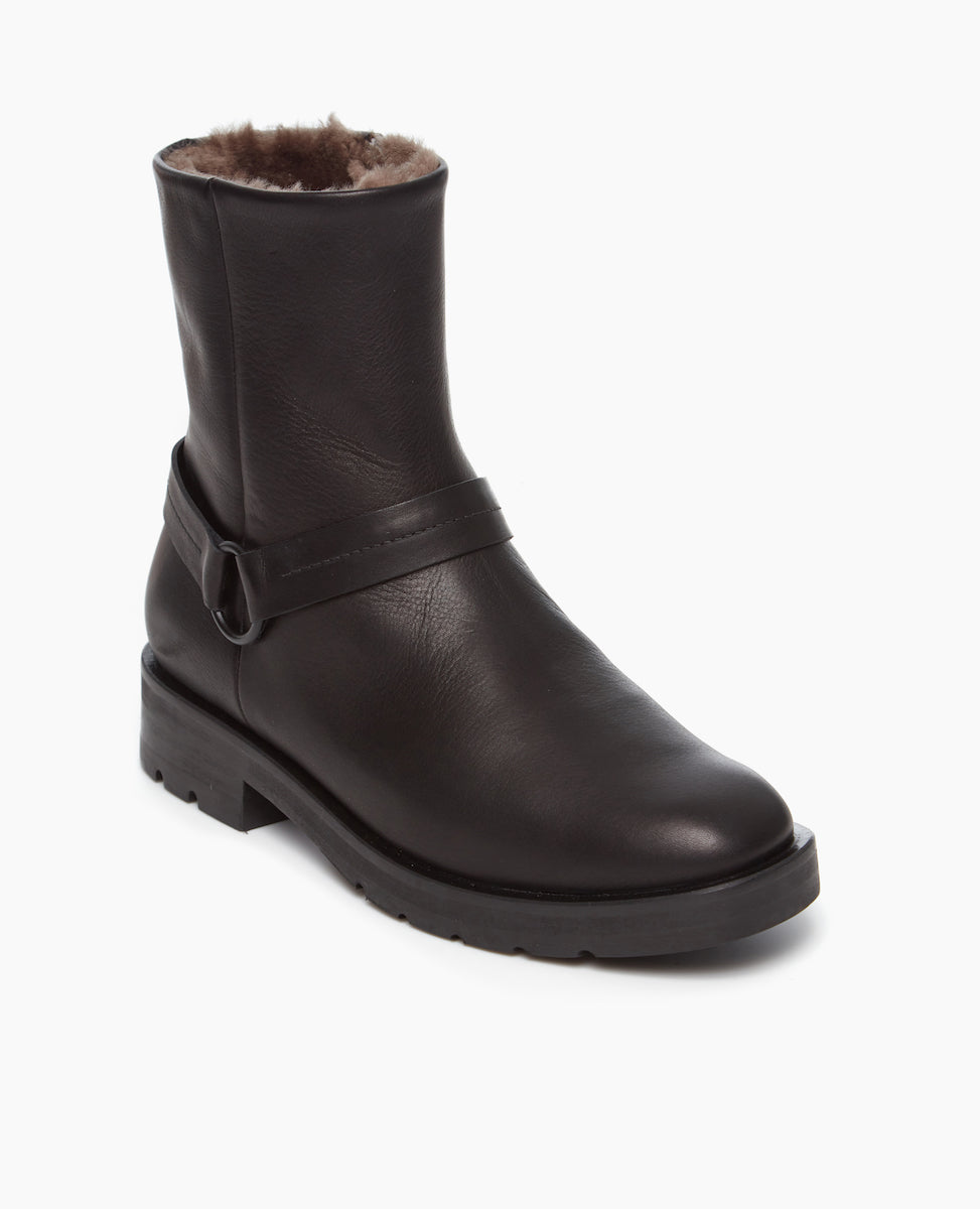 Angled view of Coclico Damiana Shearling Boot in a full shearling lined modern moto boot. This boot reatures a belt and loop at a the ankle, a lug sole, and an inside-zip closure at an angled view.