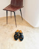 A pair of the Coclico Everly sandals on a clay floor with a rustic chair in the background. 4