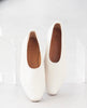 Close up of the Coclico Haloumi Flat in Greige leather: a slip-on, form-fitting flat in Italian certified sustainable leather with a squared-off toe and a 10mm solid wood heel - leaning against a clear box. 6