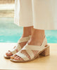 Close up angle view of model wearing Coclico Jade Heel in Greige leather. Open sandal with leather tube straps across foot, sling-back strap, solid wood block mid-height heel. 6