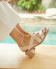 Close up of model sitting by pool wearing an open slip on sandal with soft leather straps across foot, sling-back strap, wood block mid-height heel. 2
