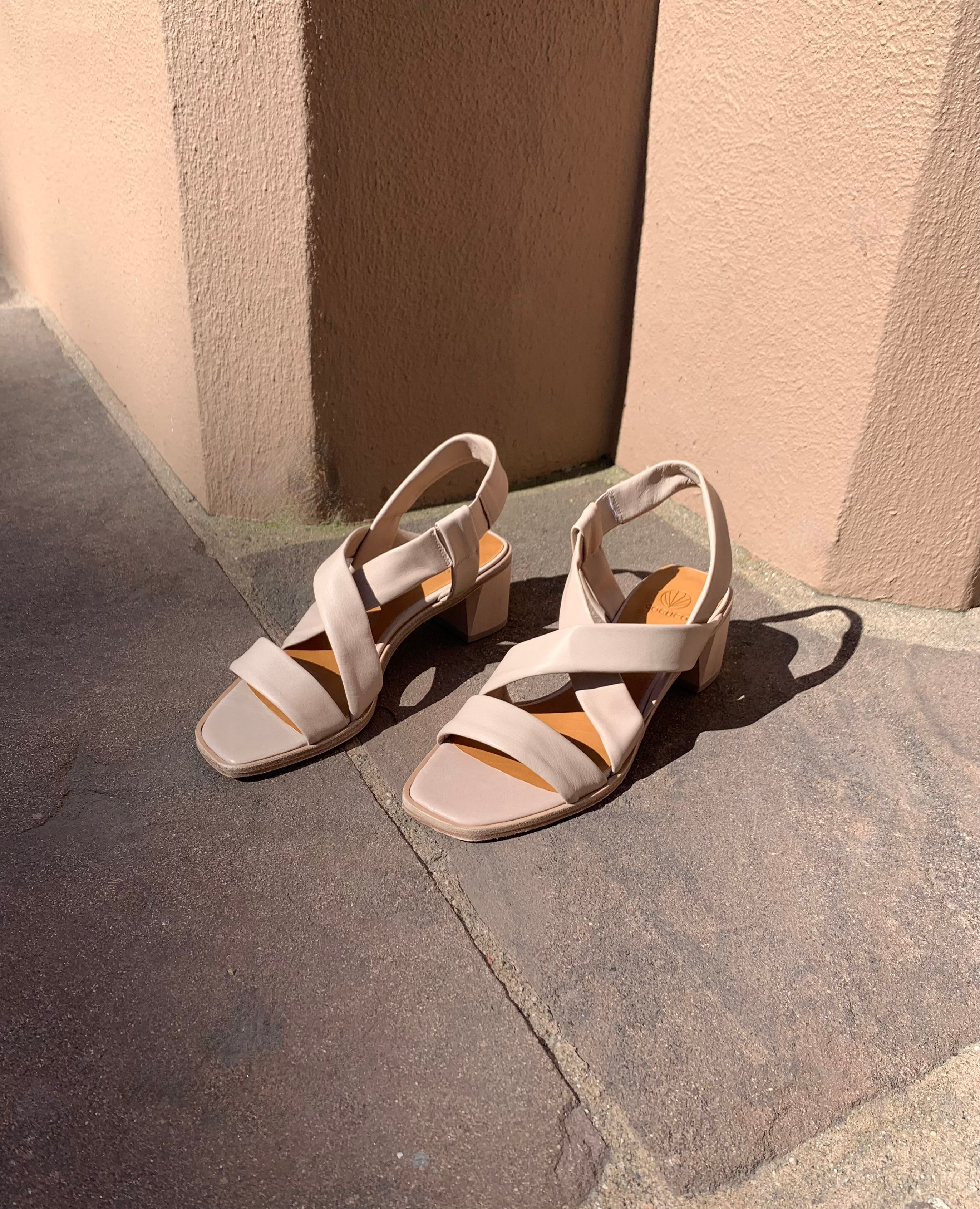 Coclico Jade Heel in Grey Mist leather an open slip on sandal with leather tube straps across the foot, sling-back strap, and wood block mid-height heel - displayed on tile flooring in sunlight.